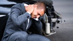 Injured man holding his neck beside a damaged car after a crash.